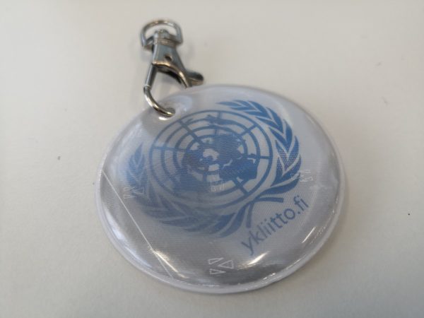 Valkoinen, pyöreä pehmoheijastin, jossa YK:n sininen maapallologo. Yläosassa metalliklipsikiinnitys