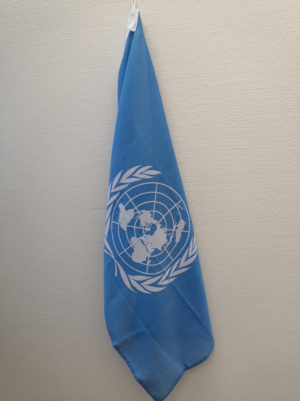 Kulmasta riippuva sininen YK:n lippu. Taustalla valkoinen seinä.