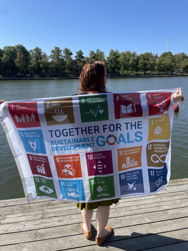 Kuvassa nainen pitelee laiturilla selässään ojennettujen käsien välissä pienempää SDG-lippua. Lipussa on valkoisella pohjalla Agenda 2030 tavoitteiden kuvakkeet ja teksti "together for the sustainable development goals"